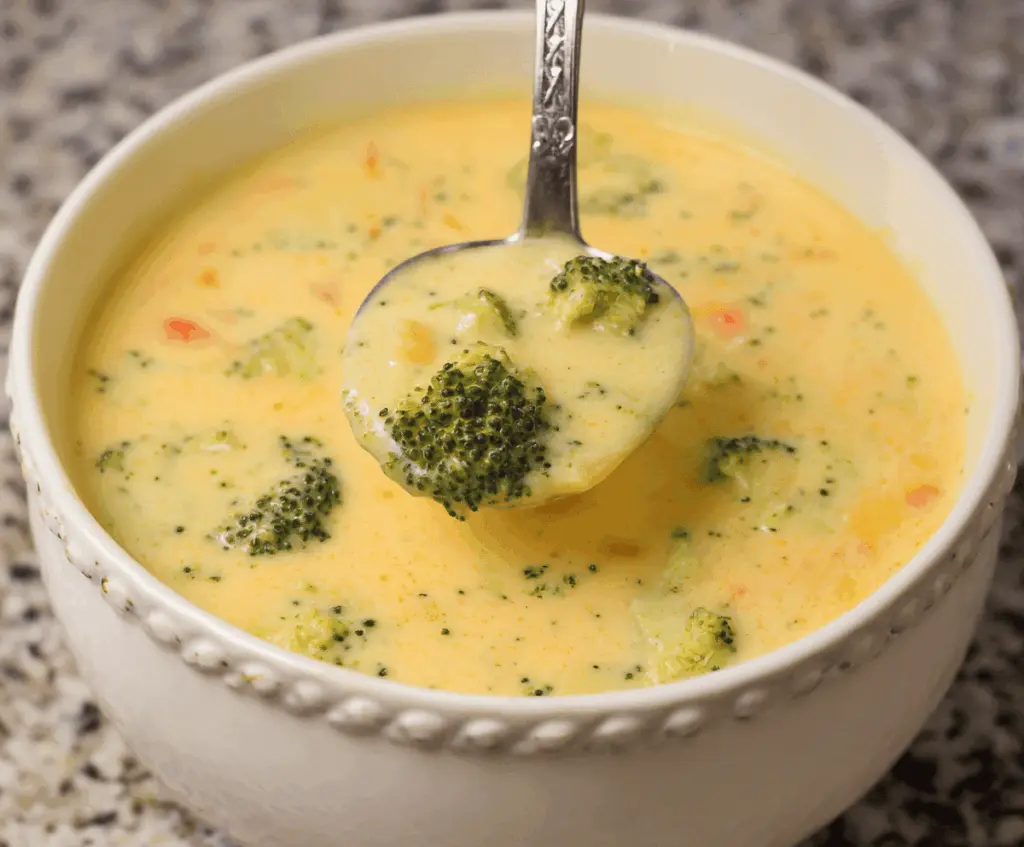 A bowl of creamy potato broccoli cheddar soup garnished with shredded cheese and fresh herbs, served with crusty bread on the side.